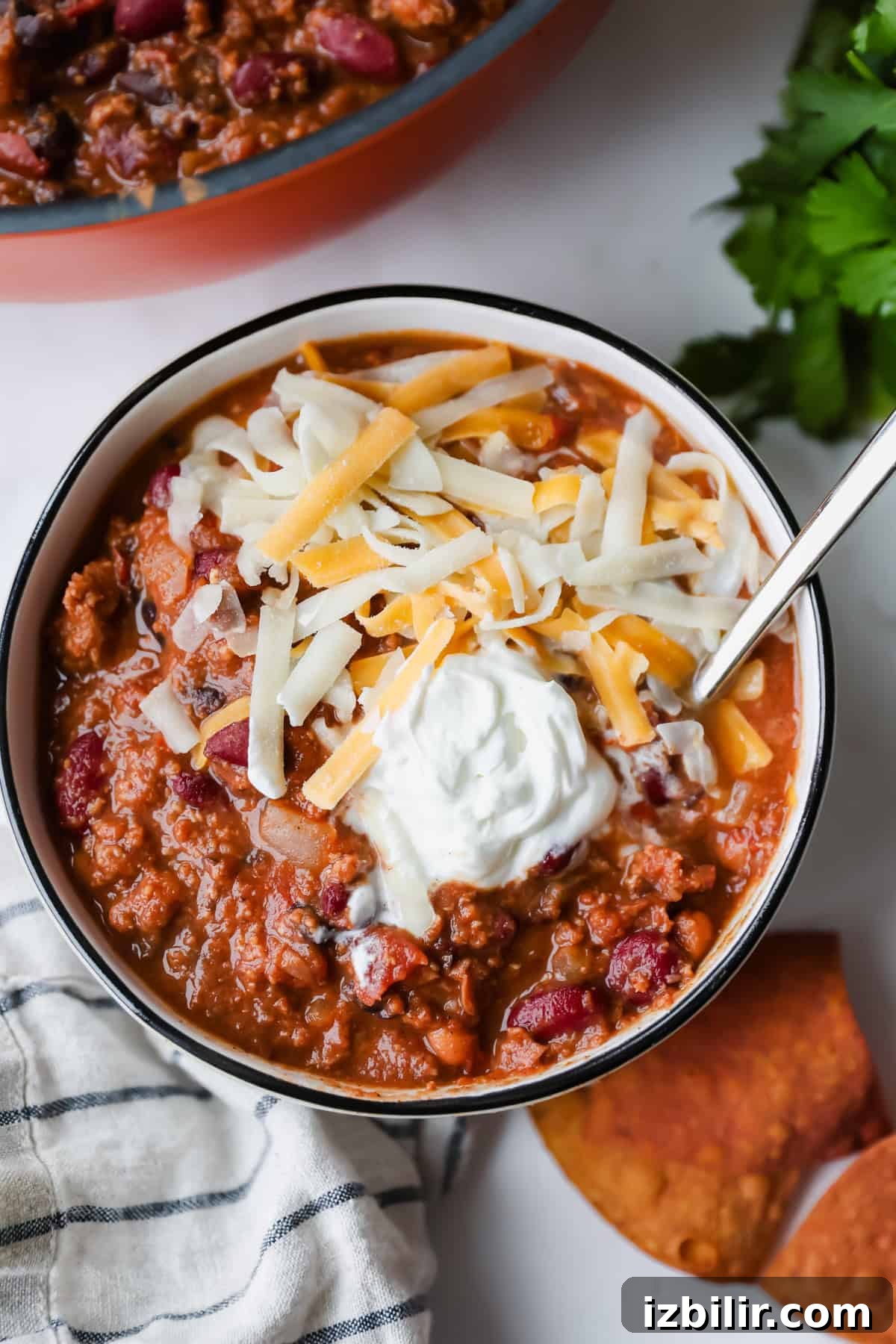 High-Protein Freezer Meals for Meal Prep 4 A bowl of chili topped with shredded cheese and a dollop of sour cream, with a spoon, tortilla chips, and a striped napkin beside it.