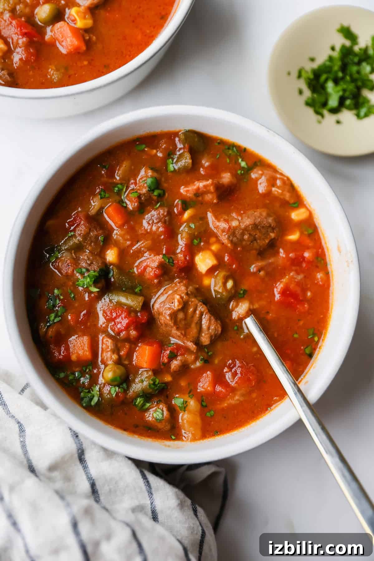 High-Protein Freezer Meals for Meal Prep 5 A bowl of beef and vegetable soup with a spoon, featuring chunks of meat, peas, carrots, corn, and tomatoes. A plate of chopped herbs sits nearby on a white surface.