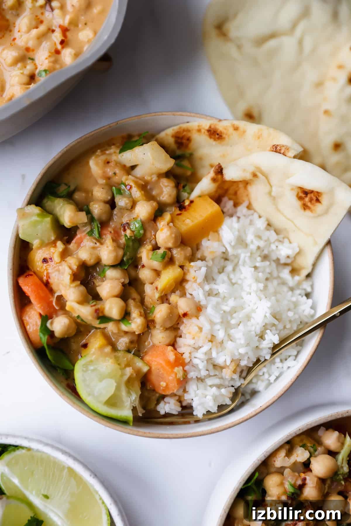 High-Protein Freezer Meals for Meal Prep 6 A bowl of chickpea curry with vegetables, white rice, lime wedge, and naan bread, served with a spoon on a white surface.