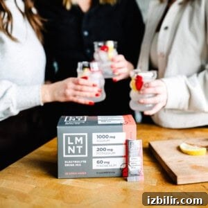 Three friends enjoying drinks, one holding a refreshing mocktail, sit beside a box of electrolyte drink mix on a wooden table.