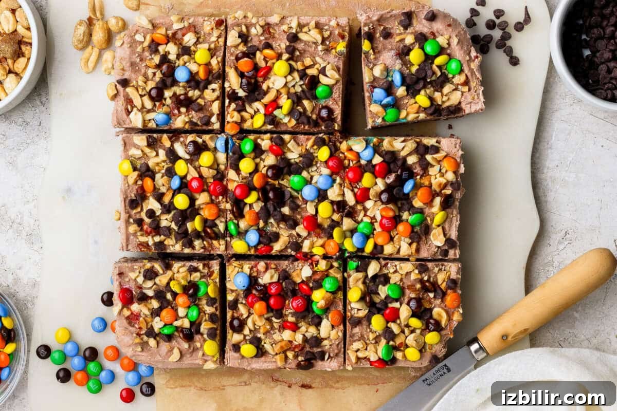 No-Bake Cottage Cheese Cheesecake Bars Recipe 2 Nine chocolate dessert bars topped with chopped peanuts, mini chocolate chips, and colorful candy-coated chocolates sit on a parchment-lined surface next to a knife and bowls of toppings.