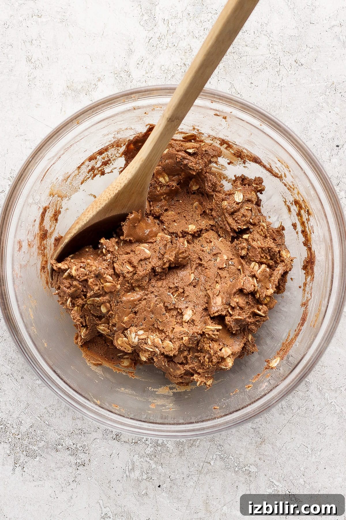 No-Bake Cottage Cheese Cheesecake Bars Recipe 3 A glass bowl contains thick chocolate cookie dough with oats, being mixed with a wooden spoon, on a light textured surface.