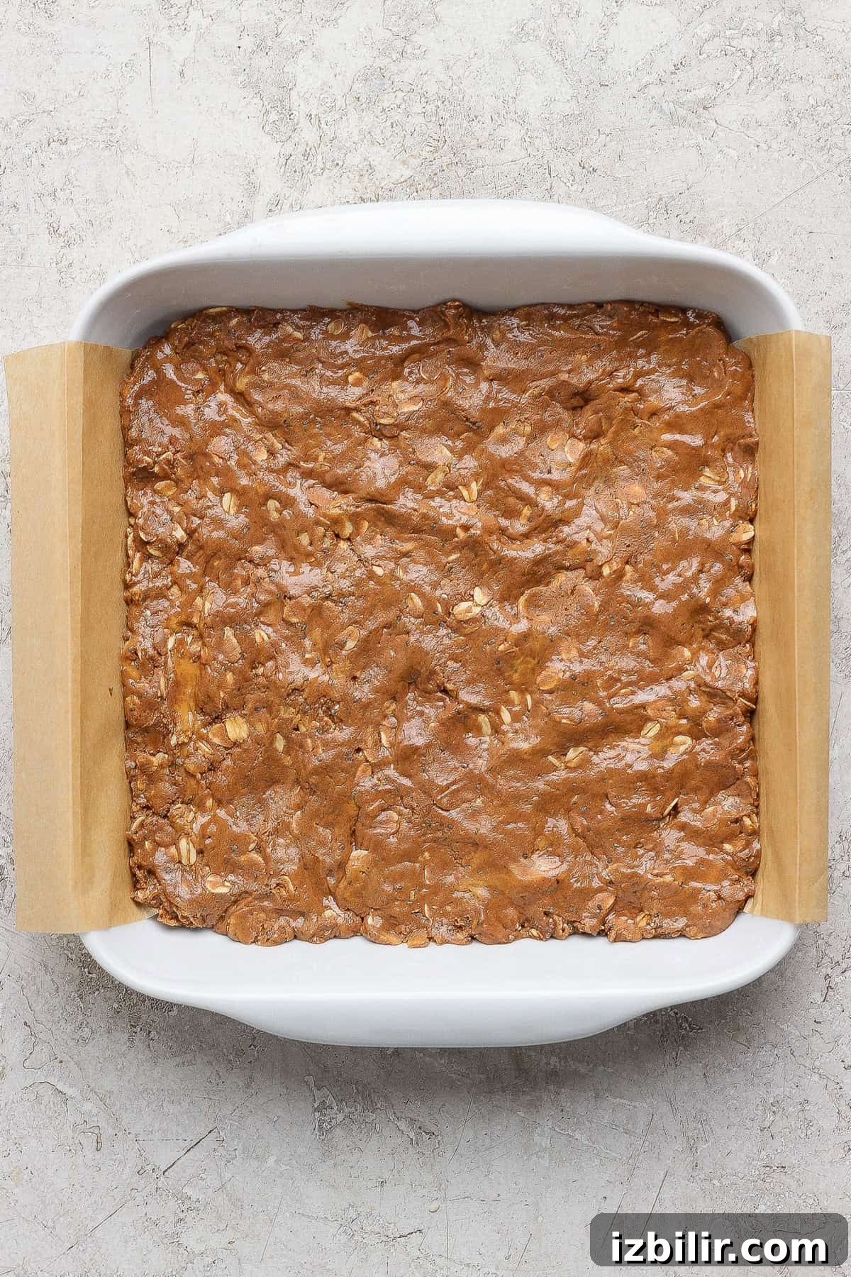 No-Bake Cottage Cheese Cheesecake Bars Recipe 6 A square white baking dish lined with parchment paper holds an uncut batch of baked oat bars on a light textured surface.