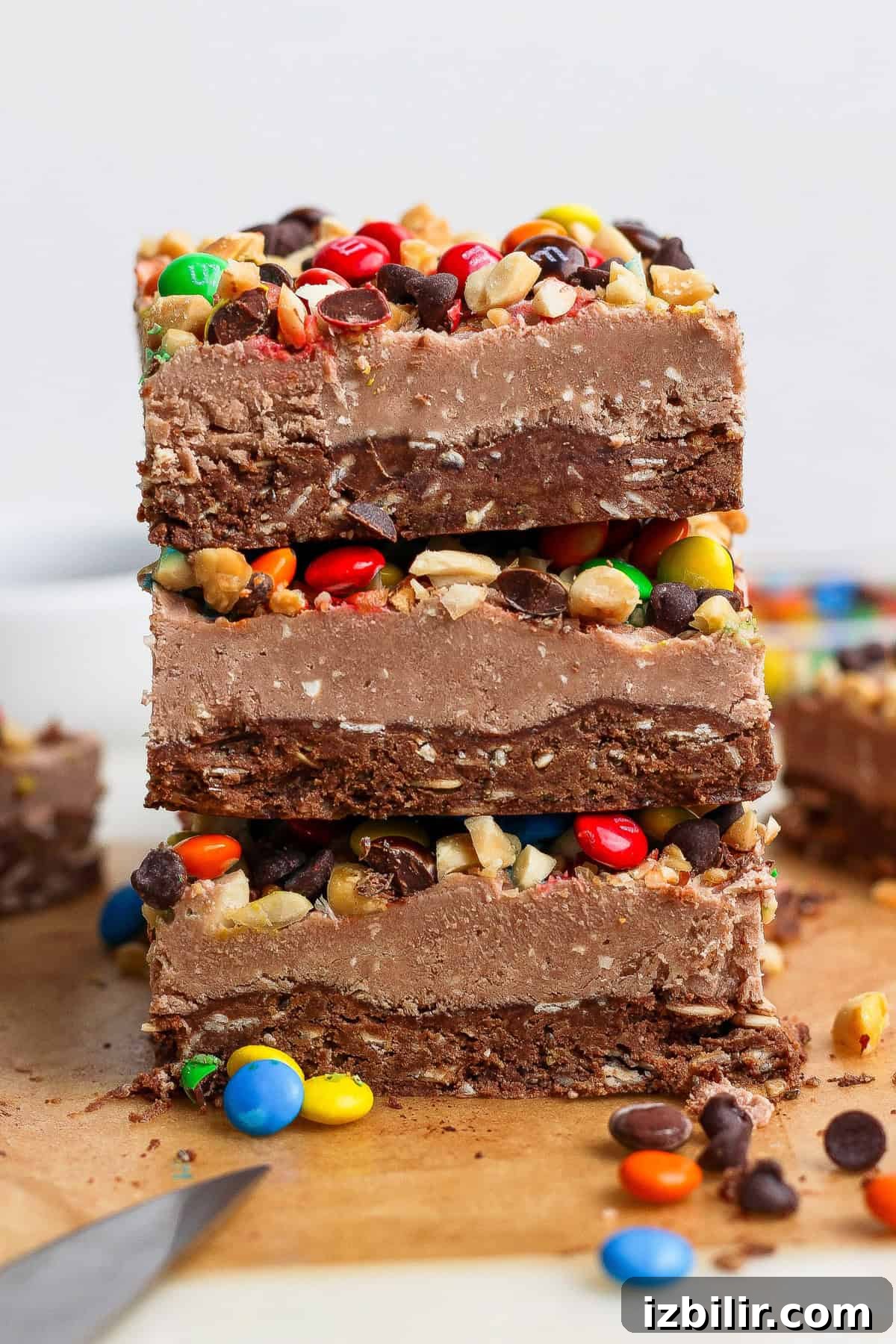 No-Bake Cottage Cheese Cheesecake Bars Recipe 7 Three stacked chocolate fudge bars topped with chopped nuts, mini chocolate candies, and chocolate chips on a wooden surface, with scattered candies around.