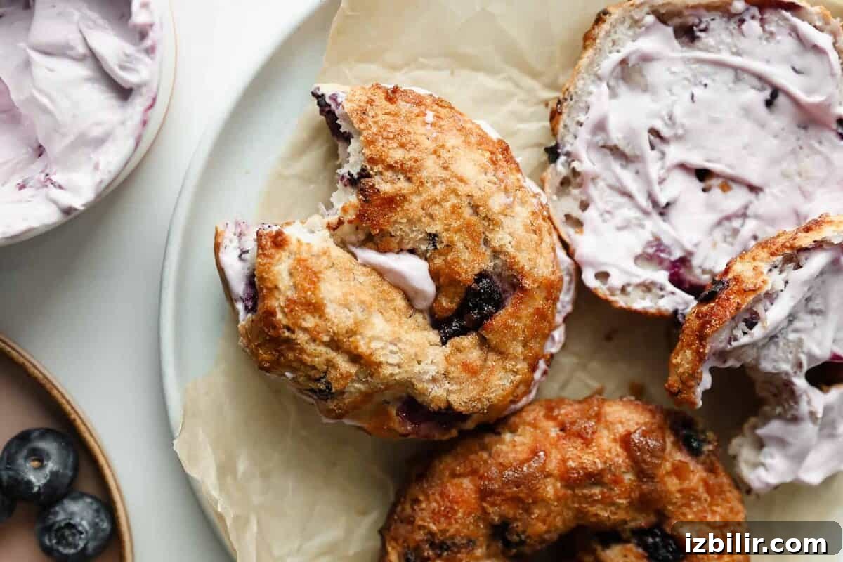 Blueberry Crunch Protein Bagels Recipe 2 A blueberry bagel with a bite taken out, filled with lavender-colored cream cheese, rests on parchment paper beside another bagel and a small bowl of blueberries.