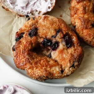 A close-up of a toasted blueberry bagel with visible blueberries, resting on parchment paper, with a partially spread bagel and purple cream cheese in the background.