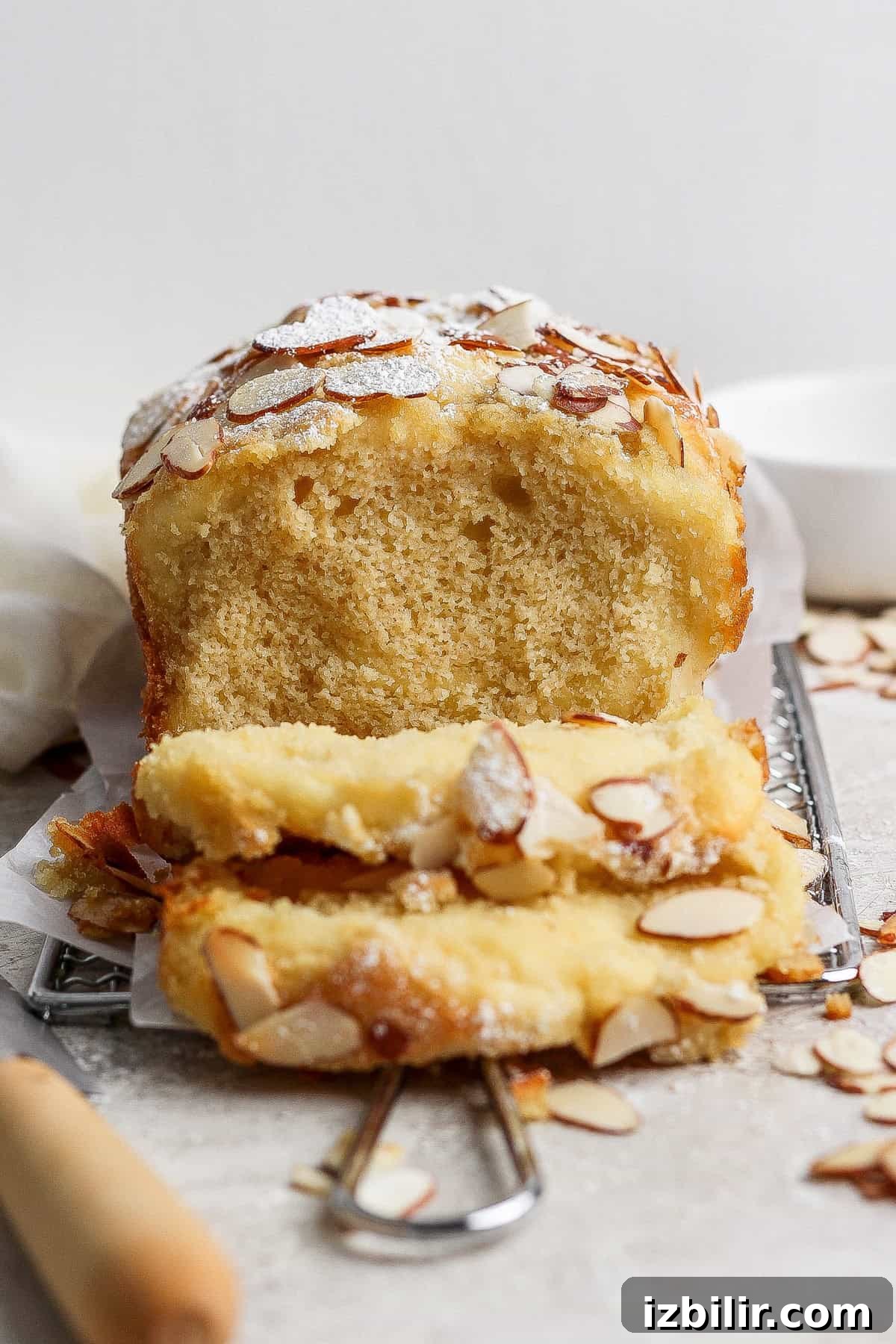 A loaf of almond cake with two slices cut, topped with sliced almonds and powdered sugar, rests on a wire rack beside a knife—evoking the cozy charm of cottage cheese pull apart bread.
