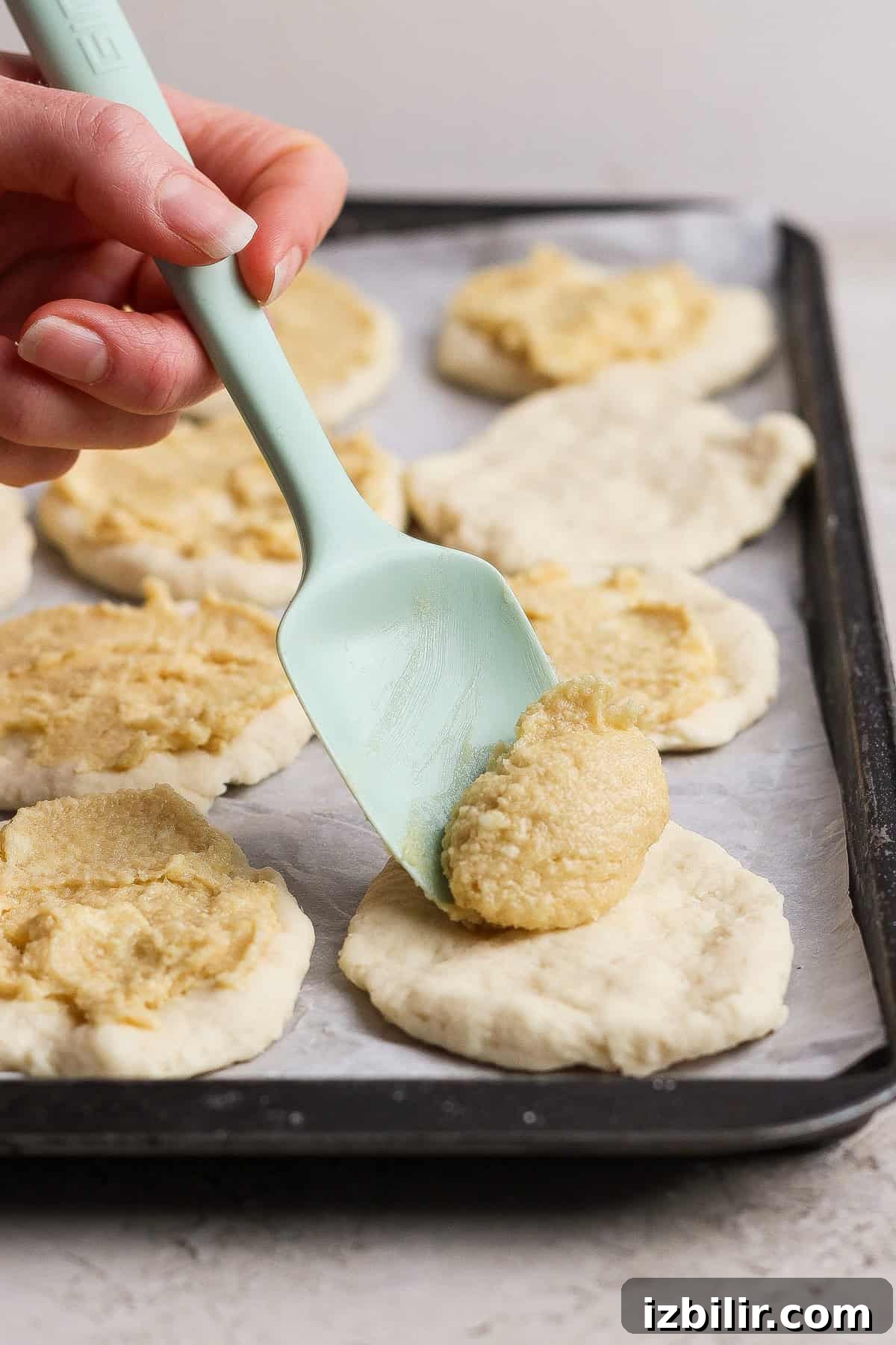 A hand uses a pale green spatula to spread cottage cheese filling onto rounds of dough for pull apart bread, all arranged on a baking sheet lined with parchment paper.