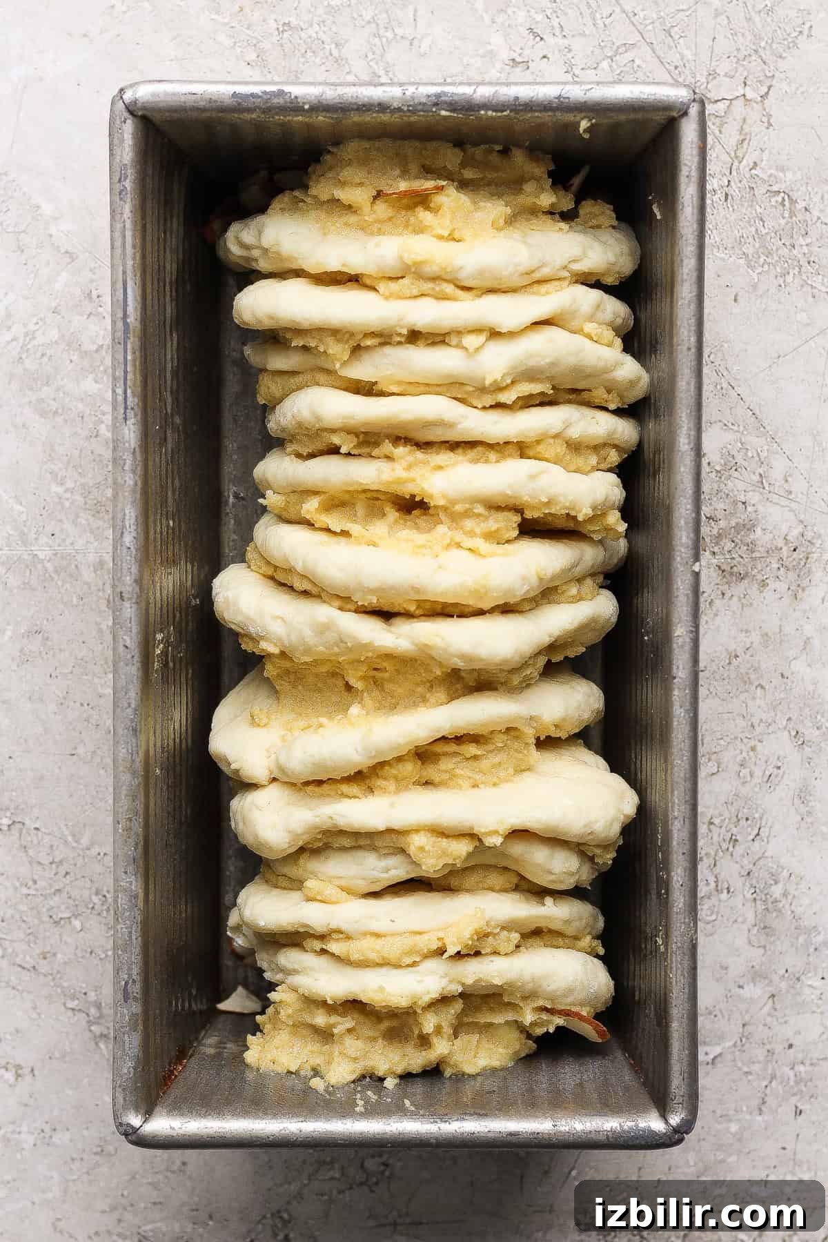 A metal loaf pan with stacked layers of unbaked dough and a yellow cottage cheese filling, arranged vertically for delicious pull apart bread, ready to bake.