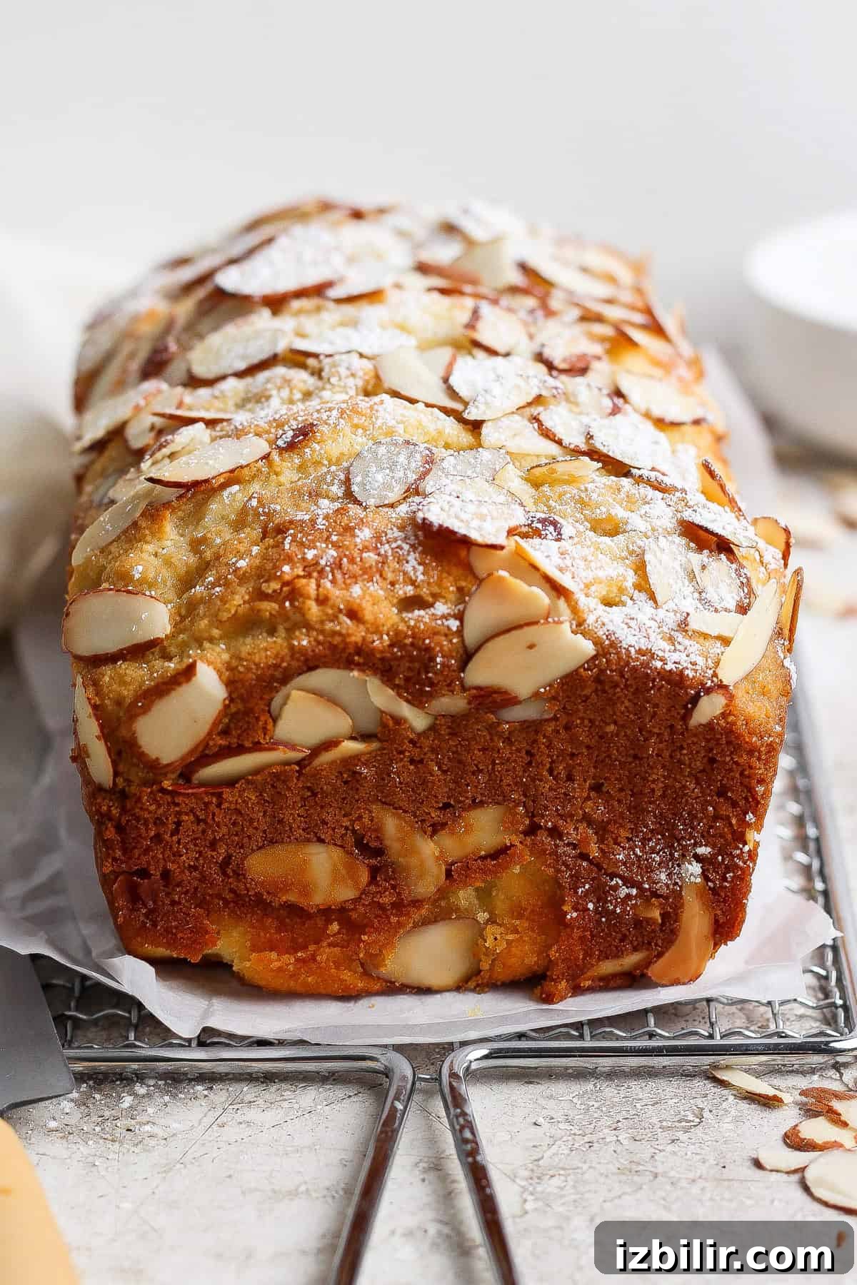A loaf of almond cake topped with sliced almonds and powdered sugar sits on parchment paper next to a cottage cheese pull apart bread on a cooling rack.