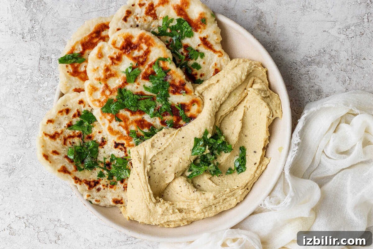 Homemade Buttery Garlic Naan Recipe with High-Protein Dough 2 A plate with grilled garlic naan flatbreads topped with chopped herbs, served beside a generous portion of hummus, on a light textured background.