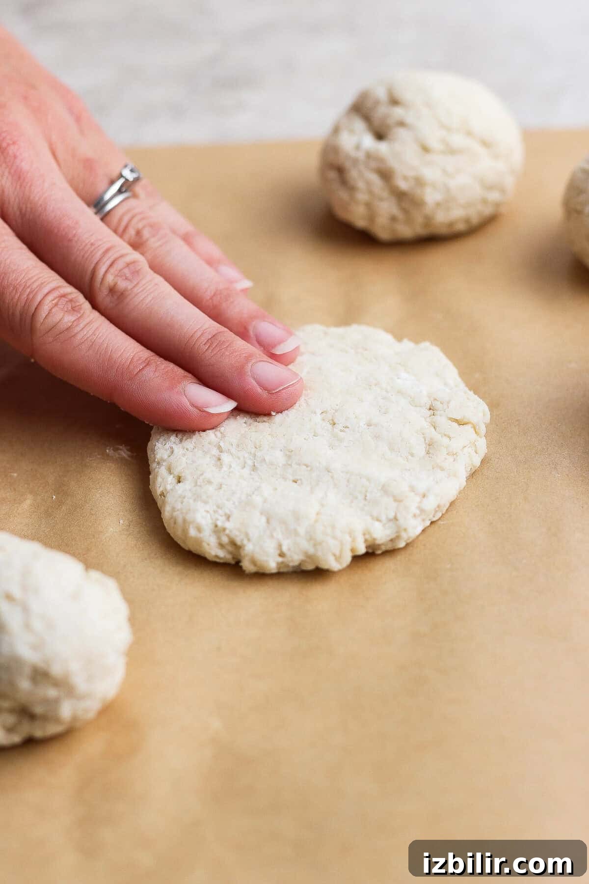 Homemade Buttery Garlic Naan Recipe with High-Protein Dough 3 A hand presses a round piece of dough flat on parchment paper, shaping garlic naan, with other dough balls nearby, preparing biscuits.
