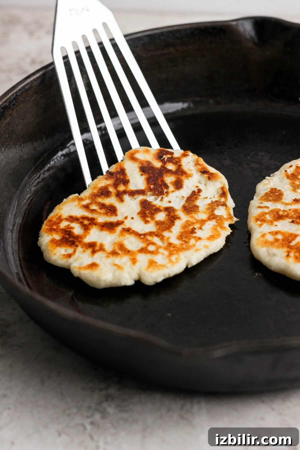 Homemade Buttery Garlic Naan Recipe with High-Protein Dough 5 A metal spatula lifts a browned garlic naan flatbread from a cast iron skillet, with another naan still in the pan.