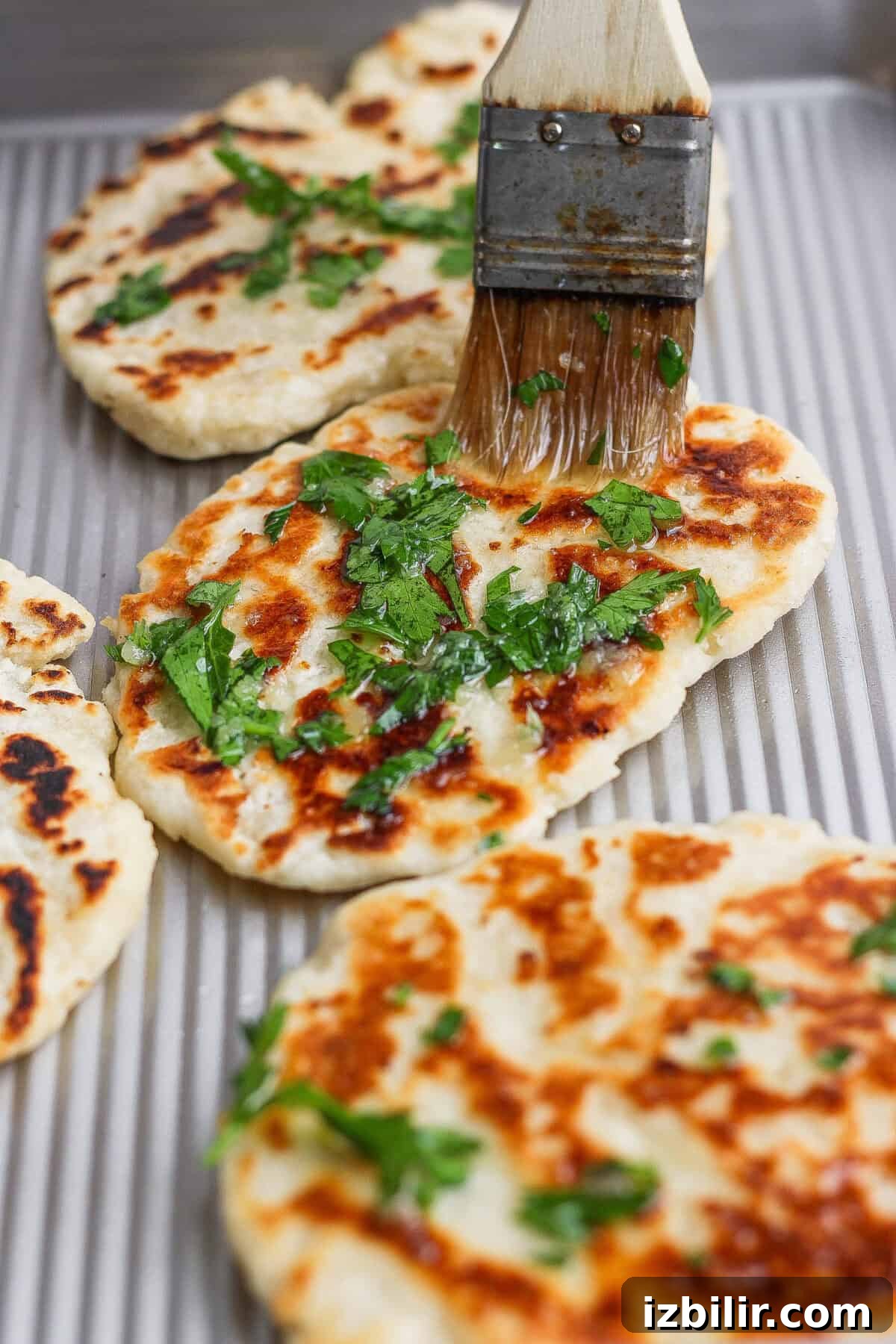 Homemade Buttery Garlic Naan Recipe with High-Protein Dough 6 A basting brush is spreading oil and chopped herbs onto freshly cooked garlic naan on a baking sheet.