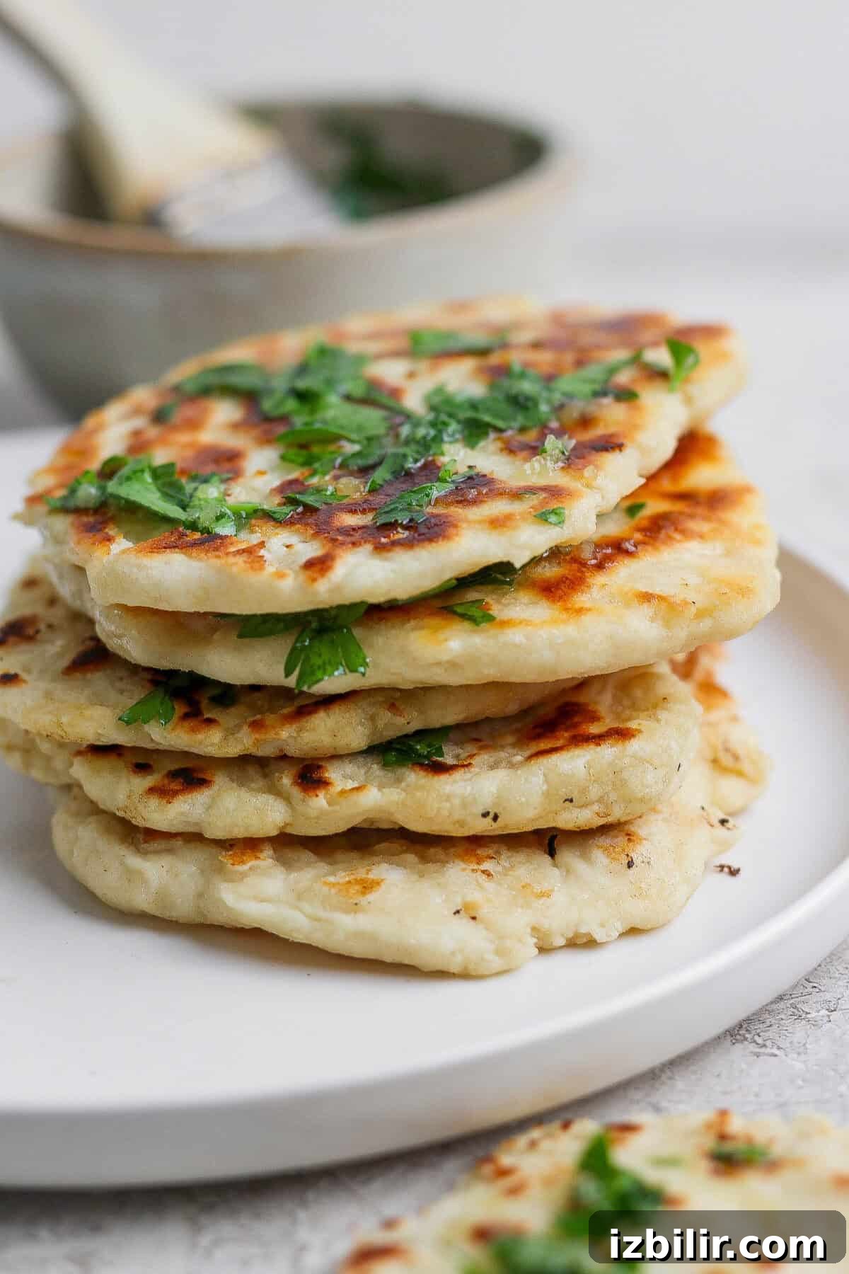 Homemade Buttery Garlic Naan Recipe with High-Protein Dough 7 A stack of golden-brown garlic naan flatbreads garnished with chopped herbs, served on a white plate.