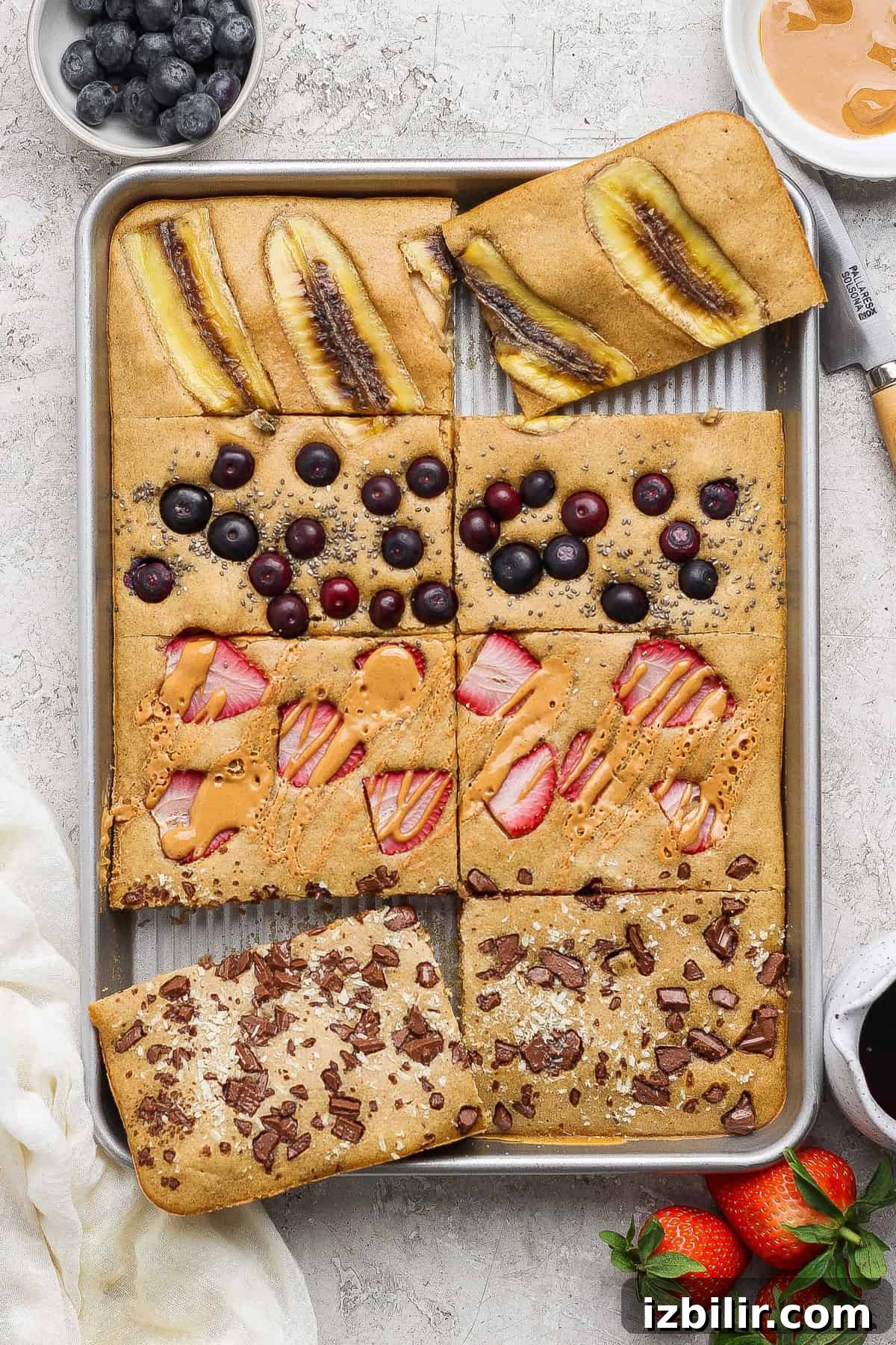 Sheet Pan Protein Pancakes Recipe - 14g Protein Per Serving 4 A baking tray with six rectangular pieces of sheet pancakes, each topped with different ingredients: banana, blueberries, strawberries with peanut butter, and chocolate chunks.