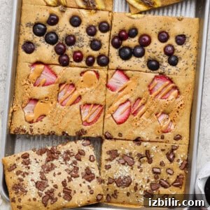 A sheet pan pancake divided into sections, each topped with different ingredients: banana, blueberries, peanut butter with strawberries, and chocolate pieces.