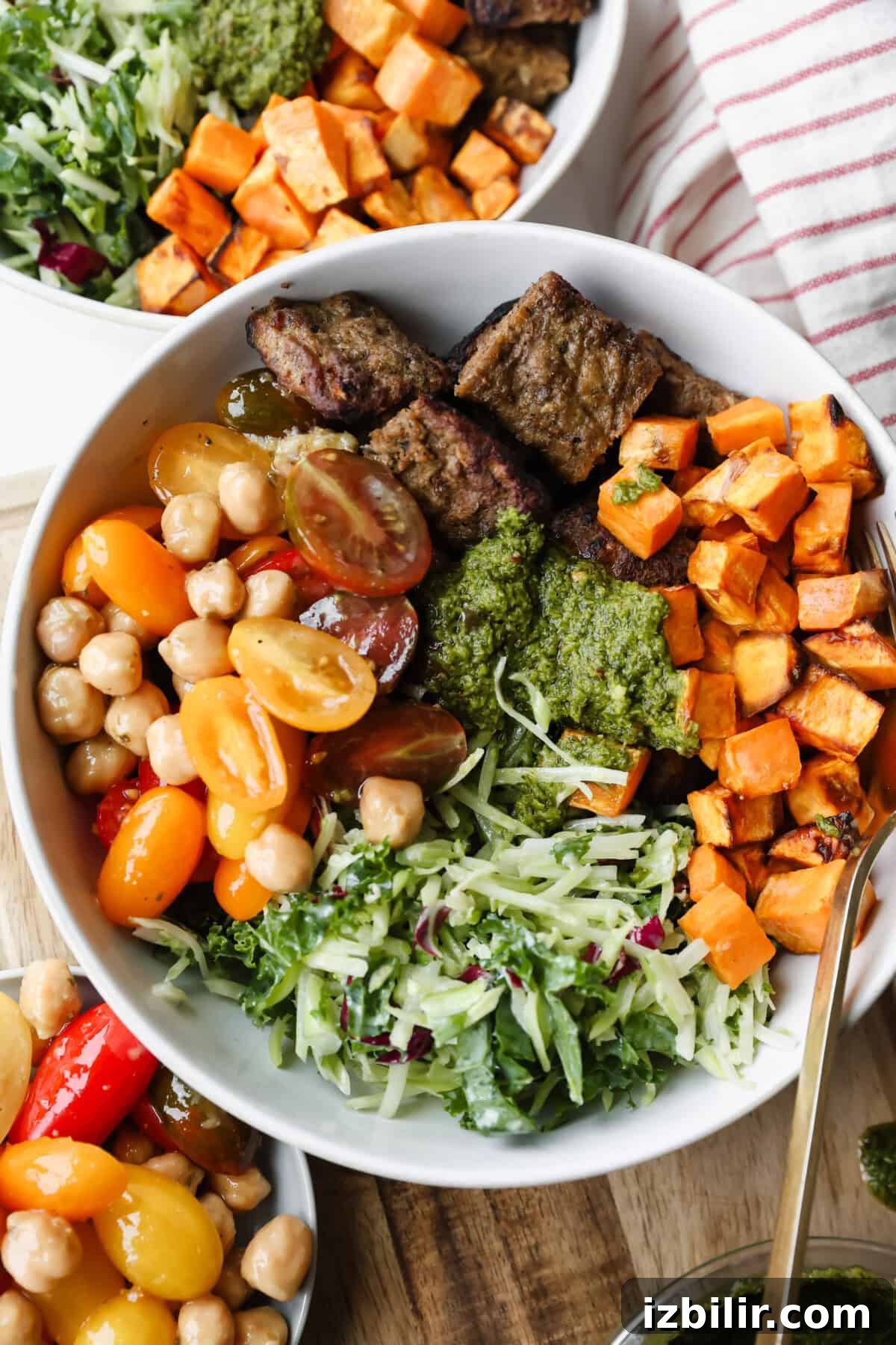A bowl filled with grilled beef strips, roasted sweet potatoes, cherry tomatoes, chickpeas, shredded greens, and green sauce, with a fork and another bowl partially visible.