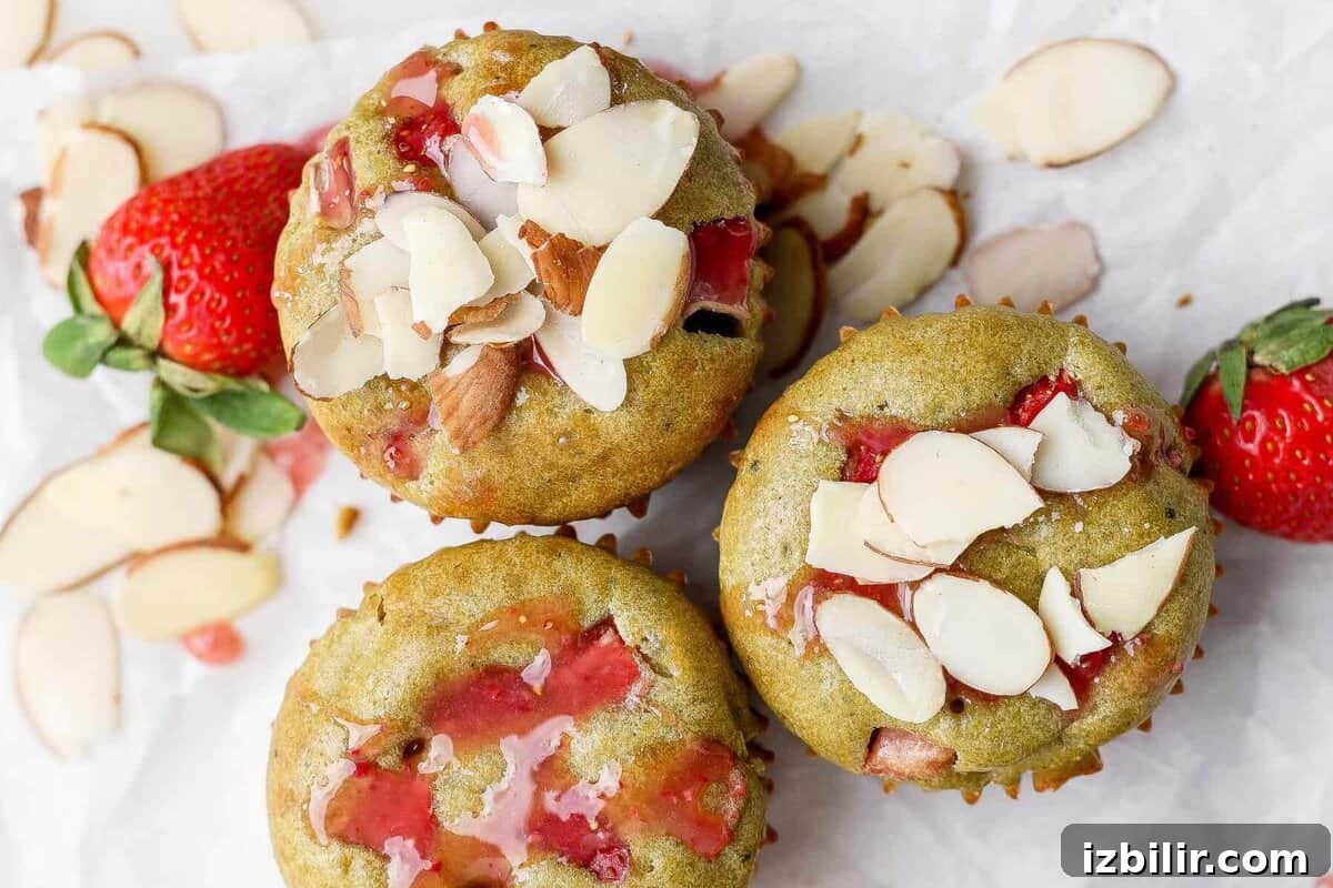 Three muffins with visible strawberry pieces and sliced almonds on top, placed on parchment paper with fresh strawberries and almond slices nearby.