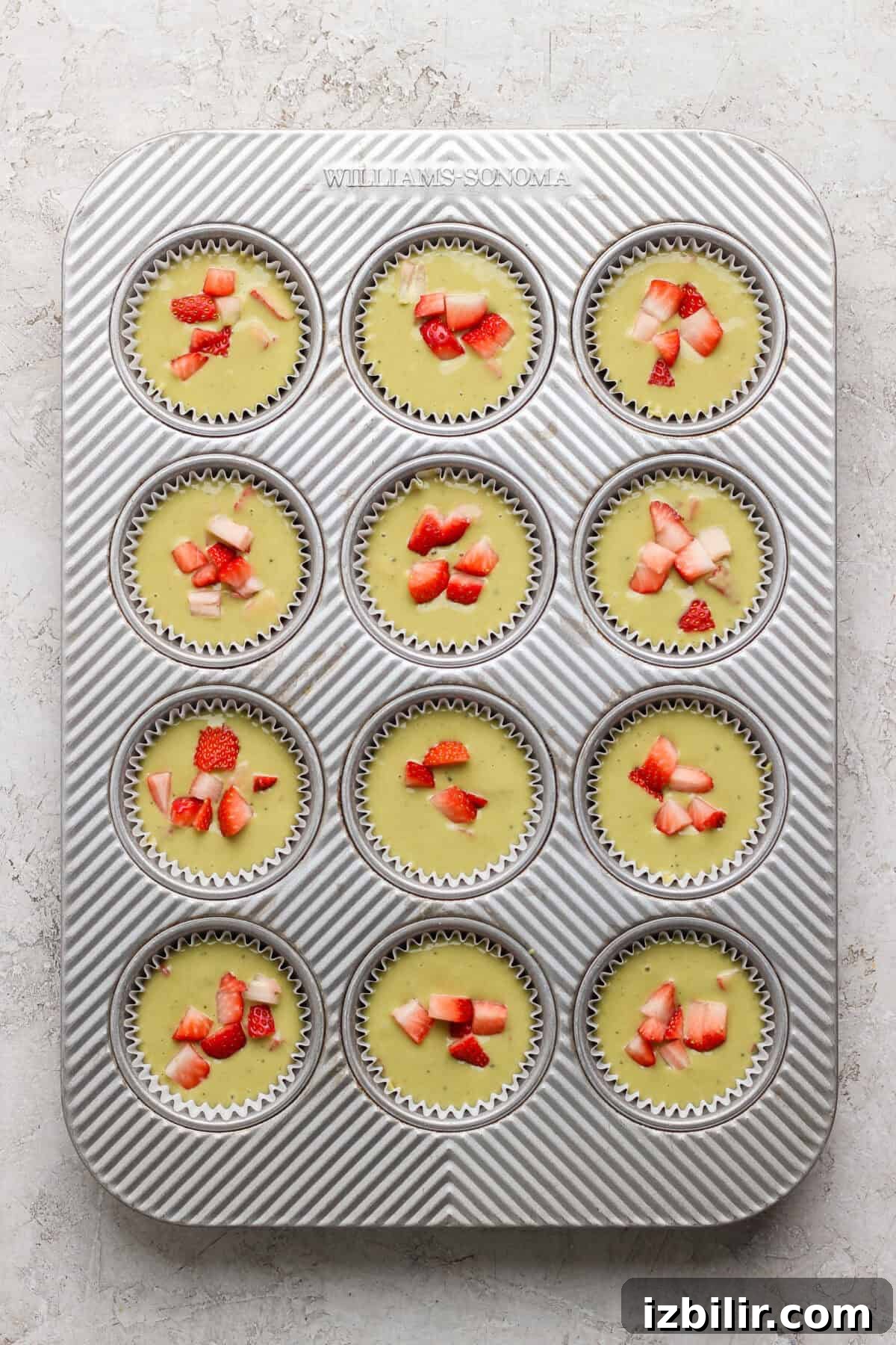 A muffin tin with twelve unbaked cupcake liners filled with batter and topped with chopped strawberries, viewed from above.