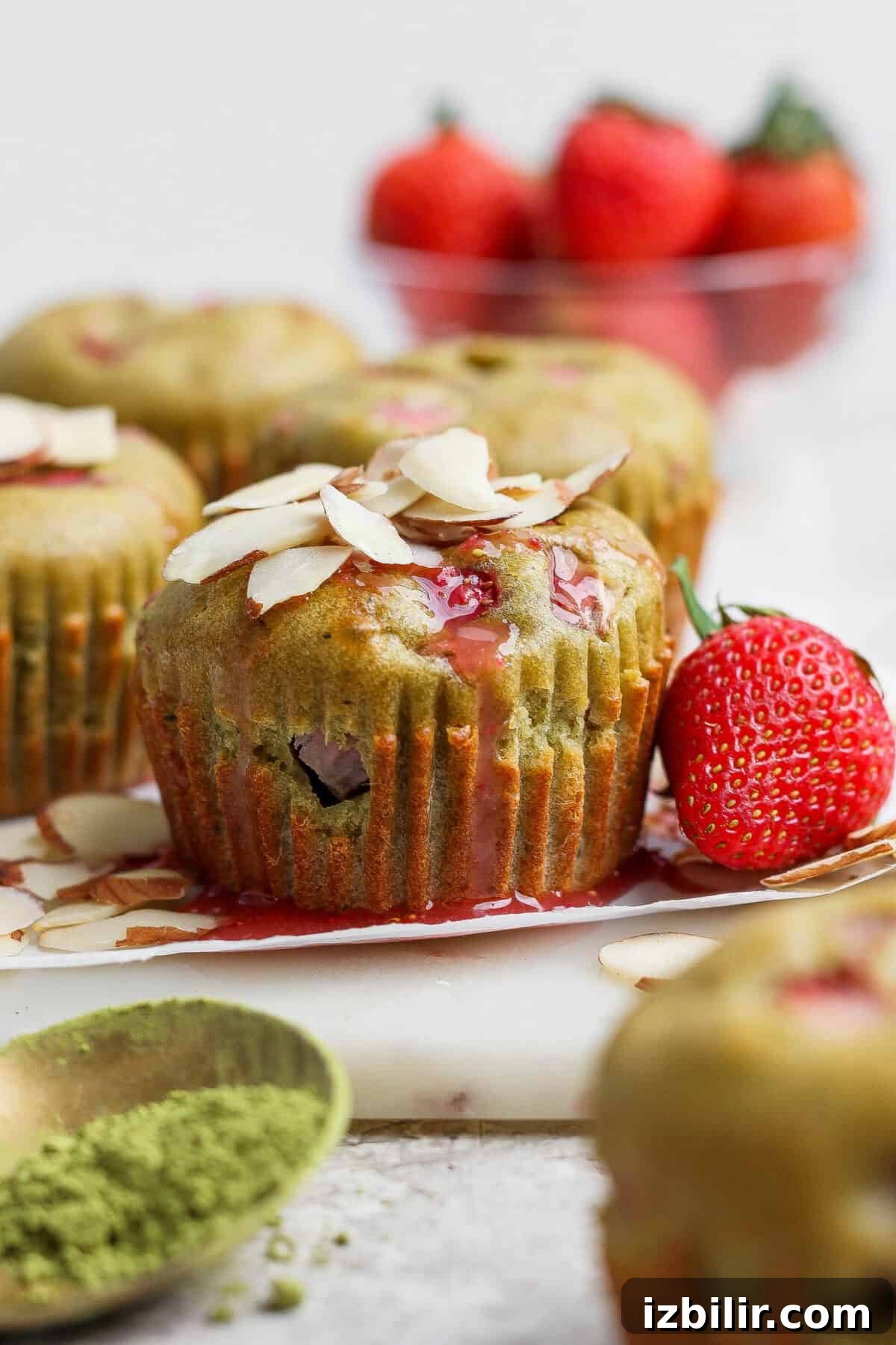 Green muffins topped with sliced almonds and bits of strawberry, with a fresh strawberry and a bowl of strawberries in the background; matcha powder in a dish nearby.