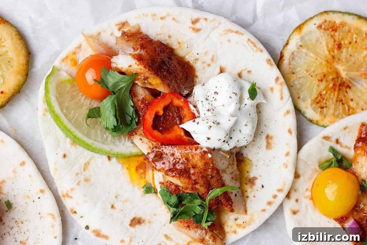 A soft flour tortilla topped with grilled fish, sliced lime, cherry tomato, red pepper, fresh parsley, and a dollop of sour cream. Grilled lime slices are visible nearby.