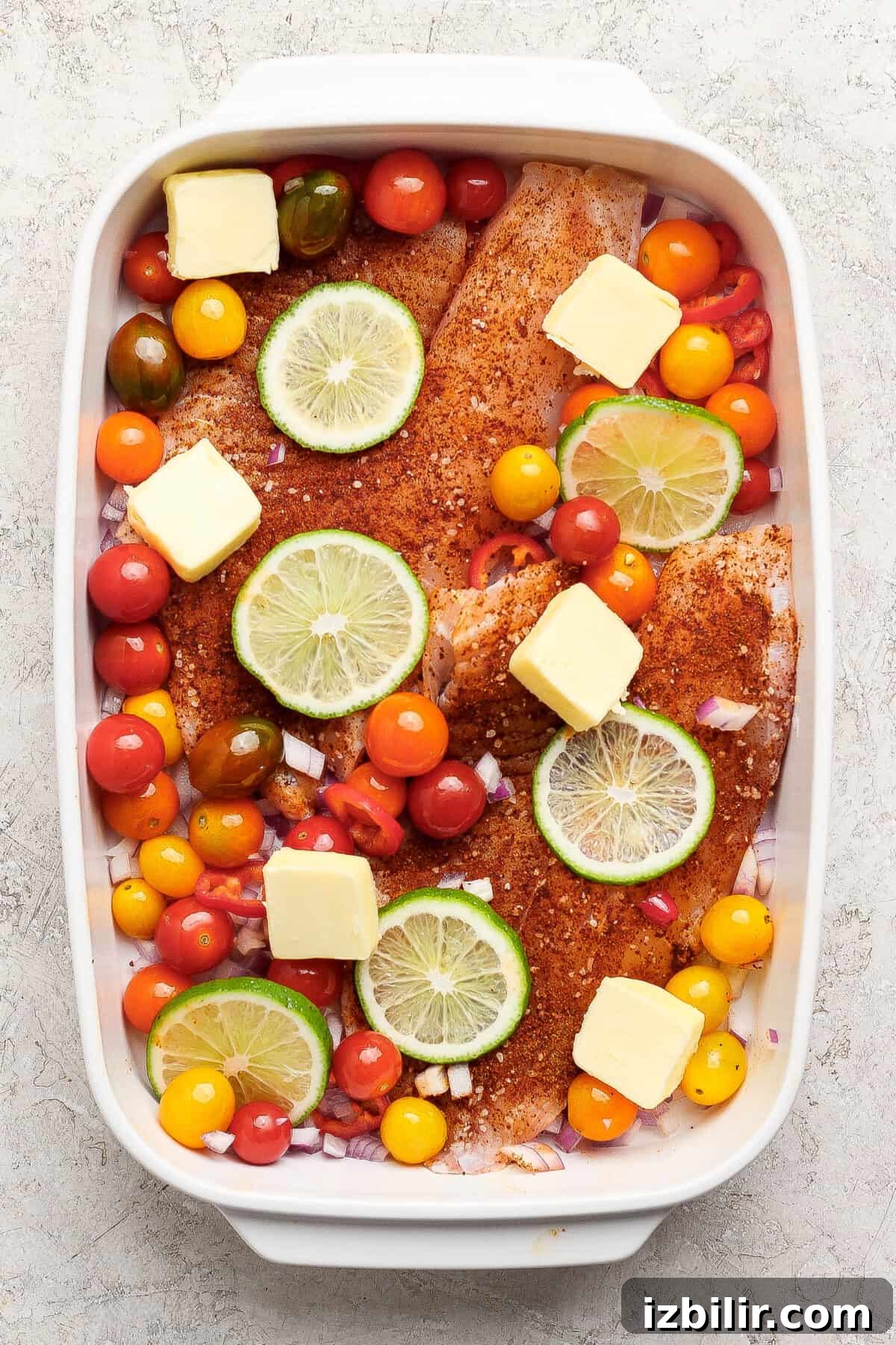 Raw seasoned fish fillets in a baking dish topped with lime slices, cherry tomatoes, onions, and butter cubes, ready for cooking.
