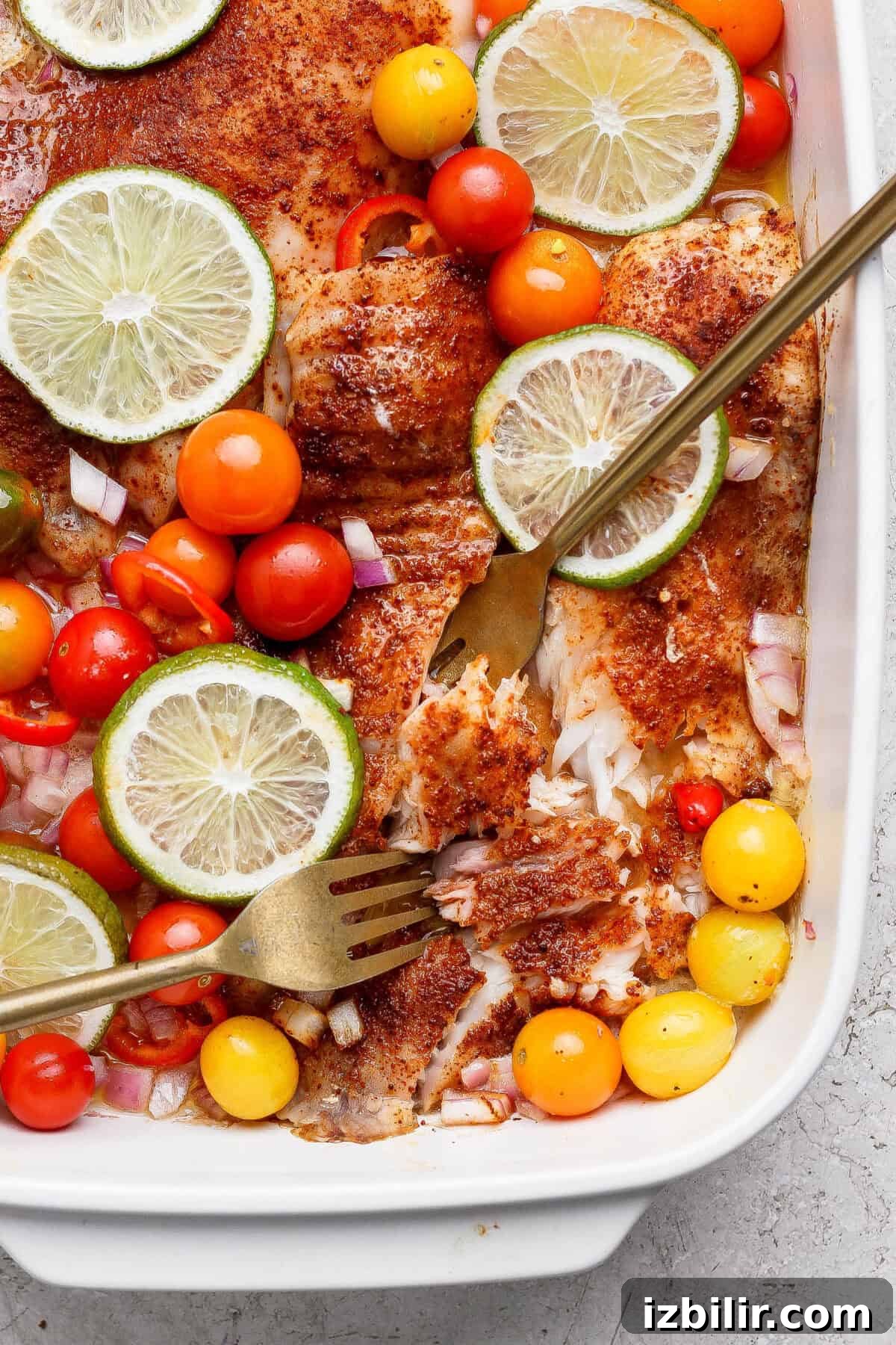 Baked fish fillets topped with sliced lime, cherry tomatoes, diced red onion, and spices, served in a white baking dish with two forks.