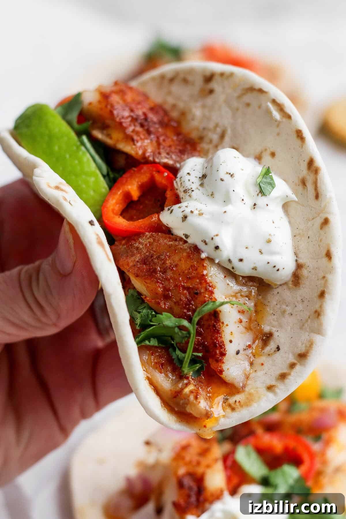 A hand holds a soft taco filled with seasoned fish, sliced red peppers, fresh greens, avocado, and a dollop of sour cream, garnished with black pepper and herbs.