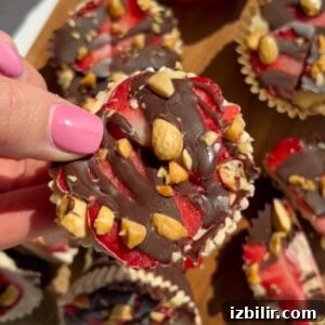 A hand with pink nail polish holds a chocolate-drizzled, nut-topped frozen treat with visible red fruit layers. More treats are arranged on a wooden surface in the background.
