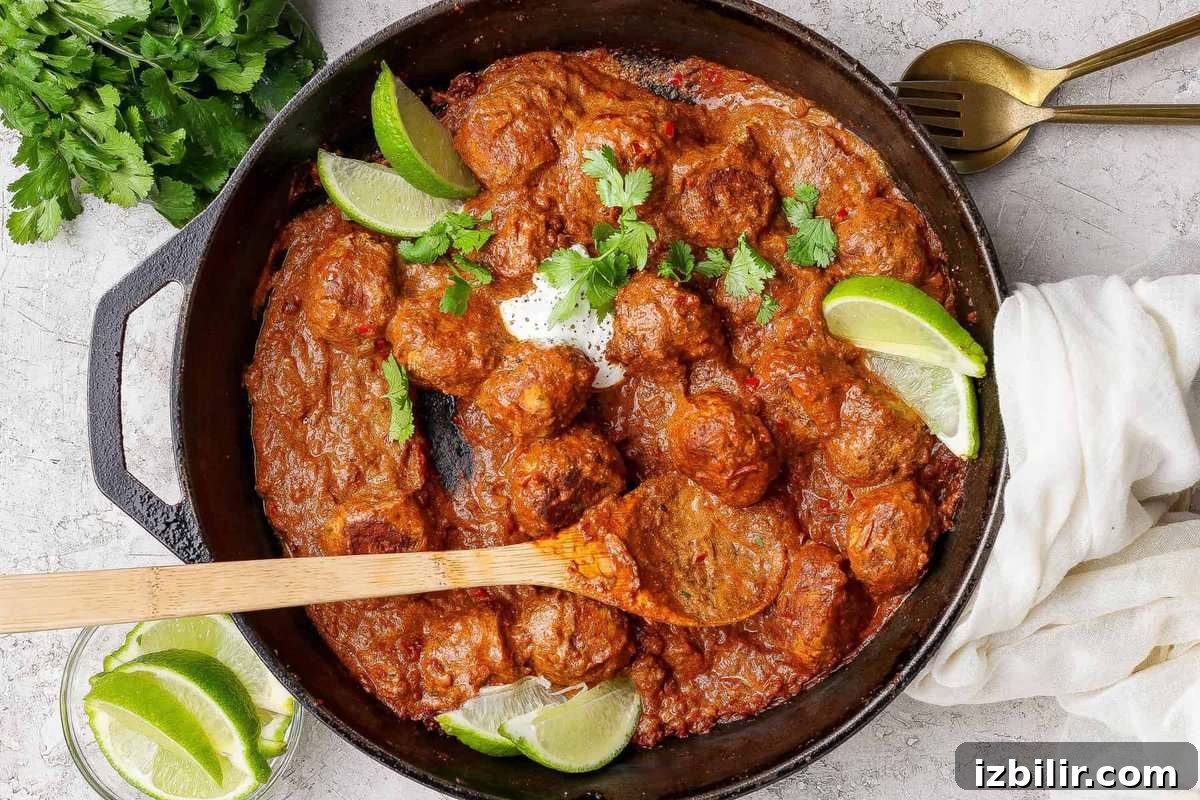 A cast iron skillet filled with butter chicken meatballs in a rich red sauce, garnished with lime wedges and cilantro, with a wooden spoon resting inside. Fresh cilantro and lime are on the side.