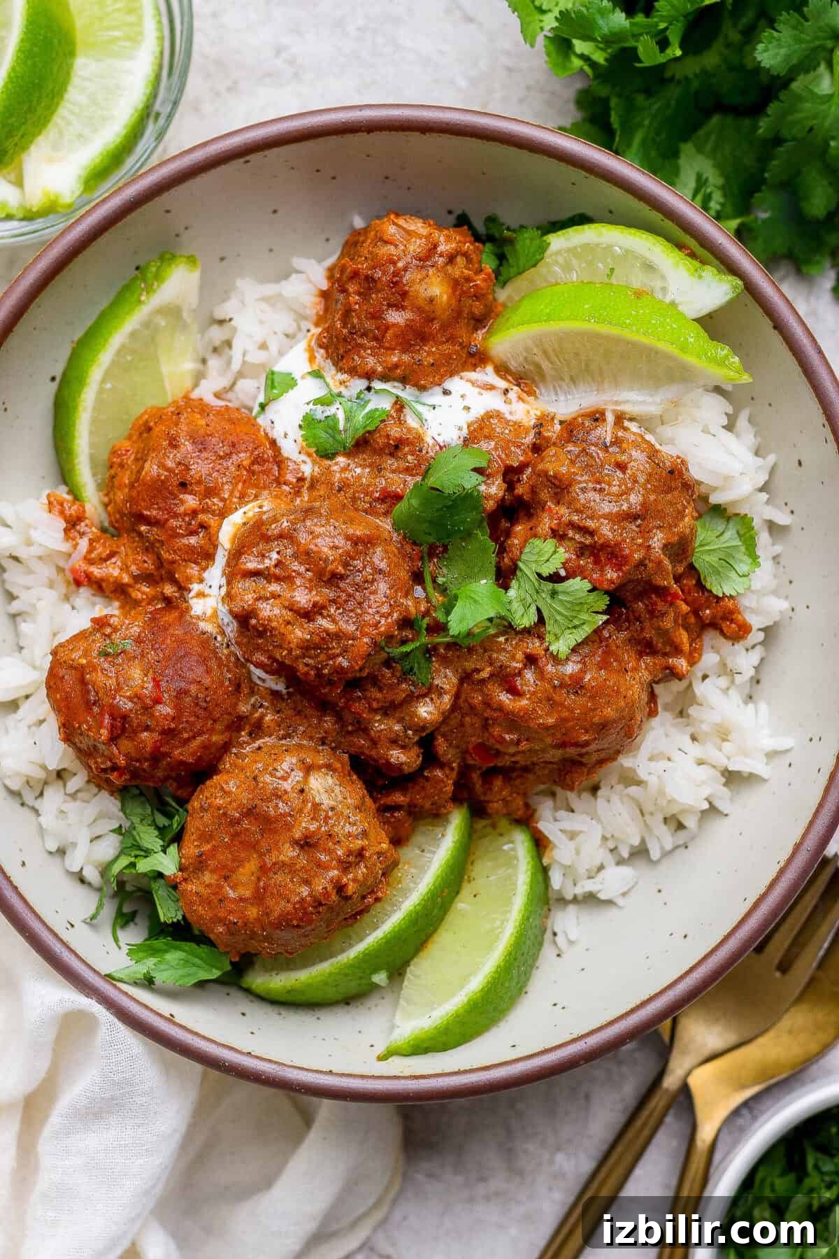 A bowl of white rice topped with butter chicken meatballs in a red sauce, garnished with lime wedges, cilantro, and a dollop of white sauce.