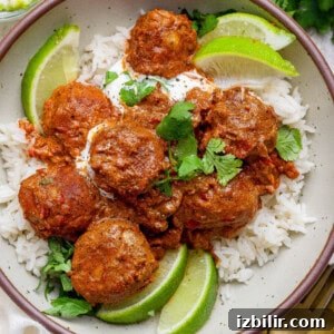 A bowl of white rice topped with butter chicken meatballs in tomato sauce, garnished with cilantro, lime wedges, and a dollop of cream.