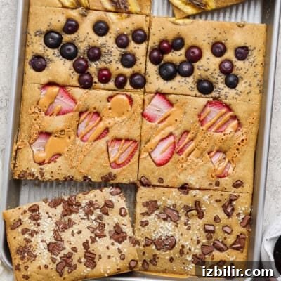 A sheet pan pancake divided into sections, each topped with different ingredients: banana, blueberries, peanut butter with strawberries, and chocolate pieces.