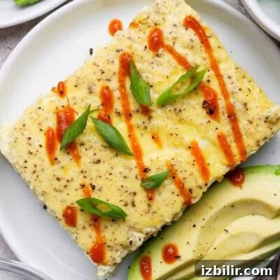 A rectangular egg dish garnished with sliced green onions and hot sauce, served with avocado slices on a white plate.
