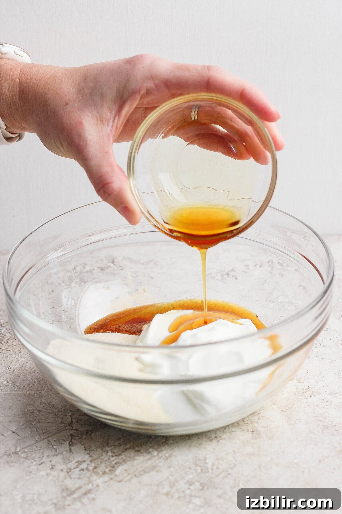 A hand pours a small glass bowl of honey into a larger bowl containing yogurt and other baking ingredients.
