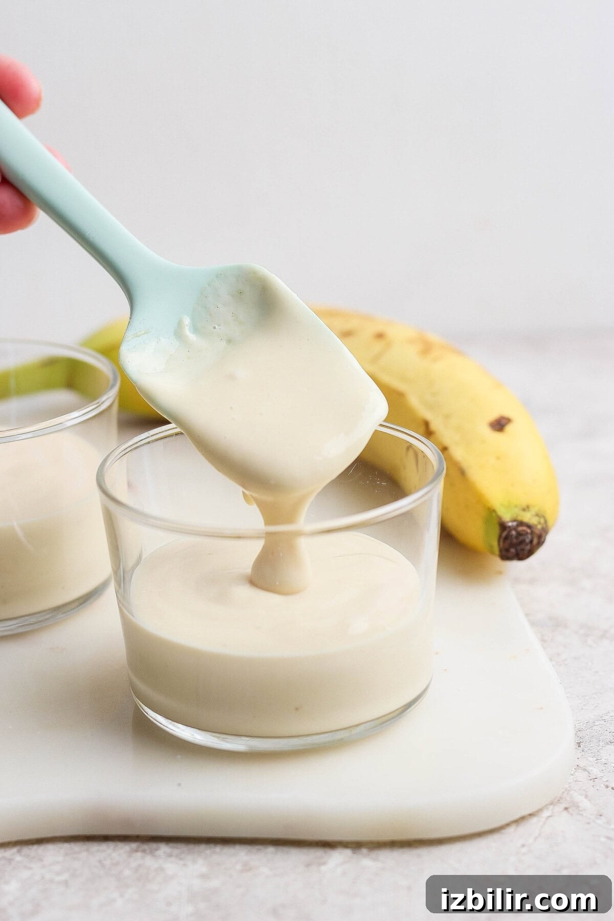 A blue spatula is lifting creamy banana pudding from a glass cup. A ripe banana and another cup of pudding are in the background on a white surface.