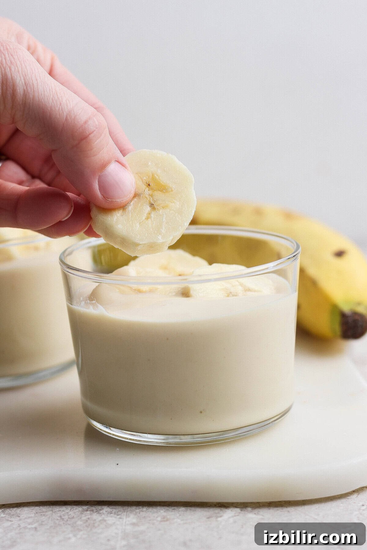 A hand placing a banana slice on top of a glass filled with banana pudding, with a whole banana in the background.
