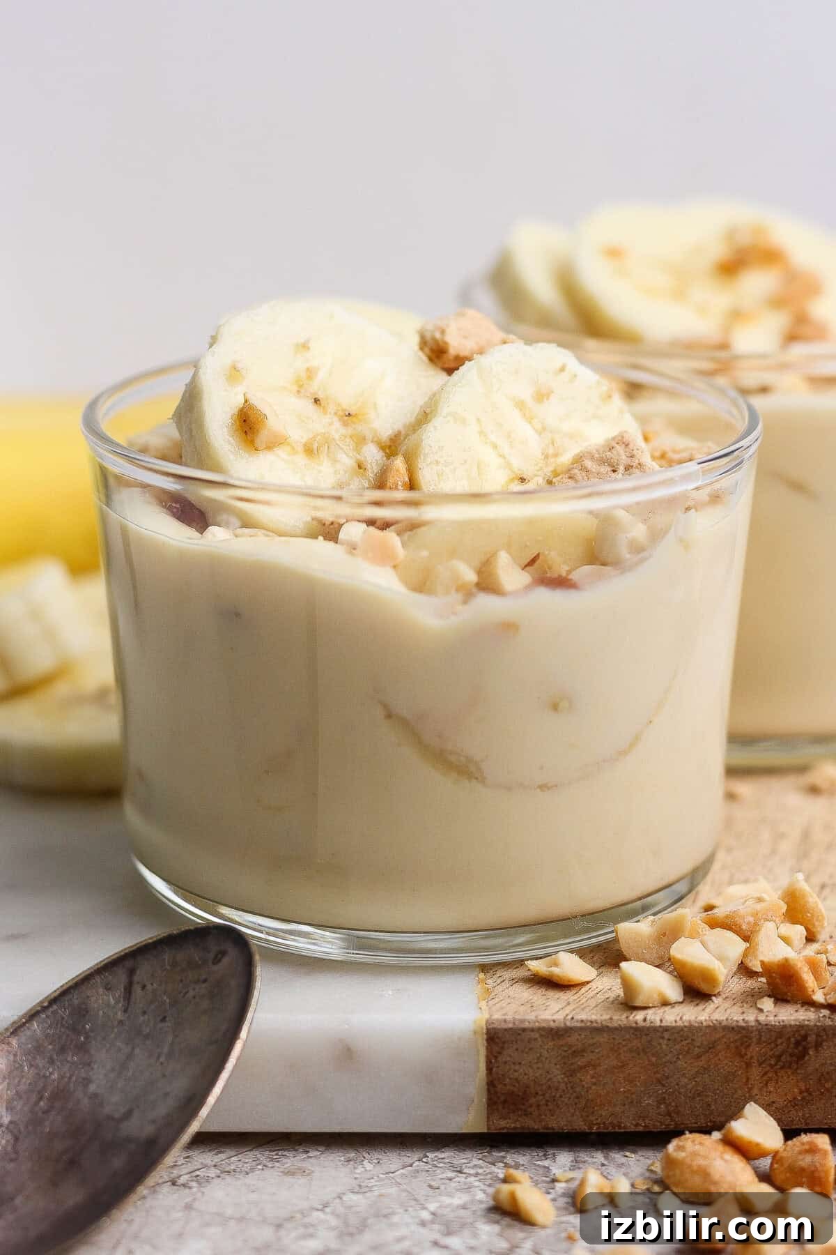 A glass cup filled with banana pudding, topped with banana slices and chopped nuts, placed on a marble surface next to a spoon and more chopped nuts.