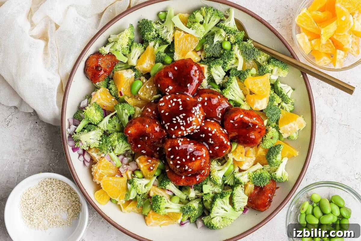 Crispy Chicken Teriyaki Recipe over Vibrant Greens 2 A bowl filled with broccoli, orange pieces, edamame, red onion, and glazed meatballs topped with sesame seeds, with small bowls of orange slices, edamame, and sesame seeds nearby.