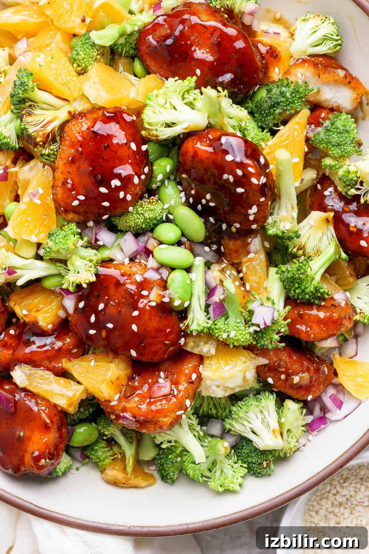 Crispy Chicken Teriyaki Recipe over Vibrant Greens 4 A bowl of glazed chicken with broccoli, orange slices, edamame, chopped red onion, and sesame seeds.