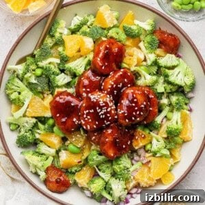A bowl of salad with broccoli, orange segments, edamame, red onion, and glazed plant-based meatballs topped with sesame seeds.