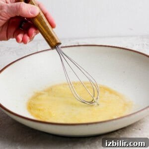 A hand holds a whisk, stirring a yellow liquid mixture in a shallow, white bowl on a light countertop.
