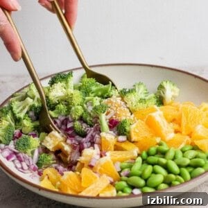 A bowl with chopped broccoli, red onion, orange segments, and edamame being mixed with two gold utensils by a person’s hands.