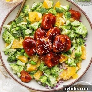 A bowl filled with broccoli, orange pieces, edamame, red onion, and glazed meatballs topped with sesame seeds, with small bowls of orange slices, edamame, and sesame seeds nearby.