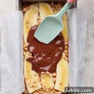 A hand uses a spatula to spread melted chocolate over sliced bananas and chopped nuts in a parchment-lined loaf pan.