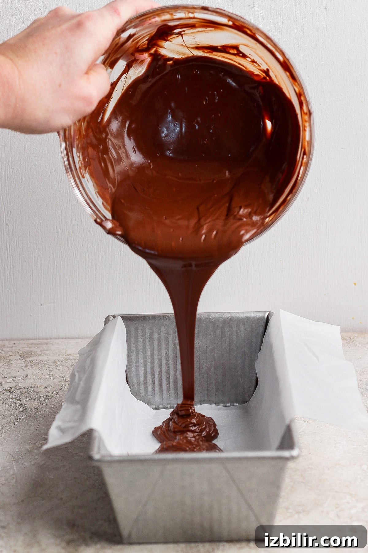 A person pours thick melted chocolate from a glass bowl into a parchment-lined loaf pan on a light-colored surface.