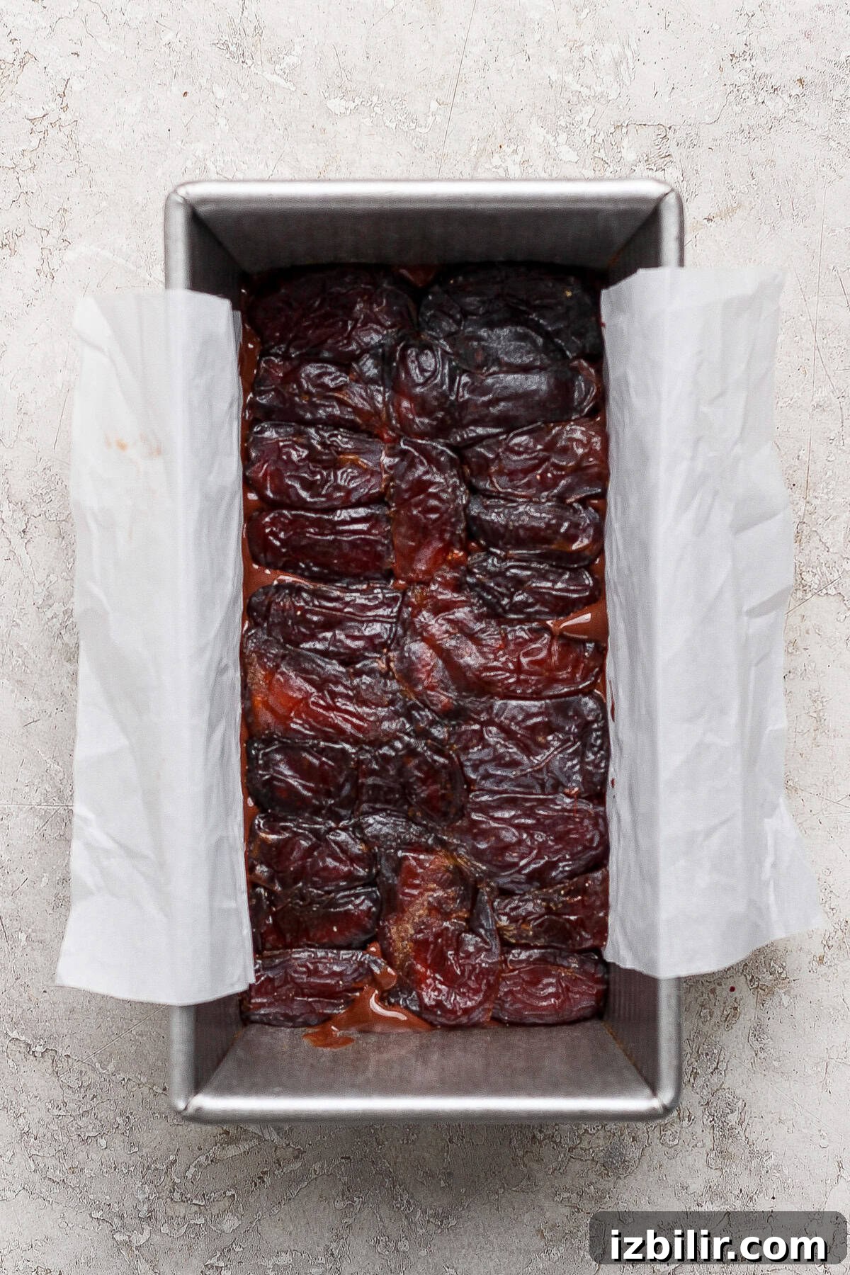 A rectangular metal baking pan lined with parchment paper, filled with a layer of pitted dates arranged closely together.