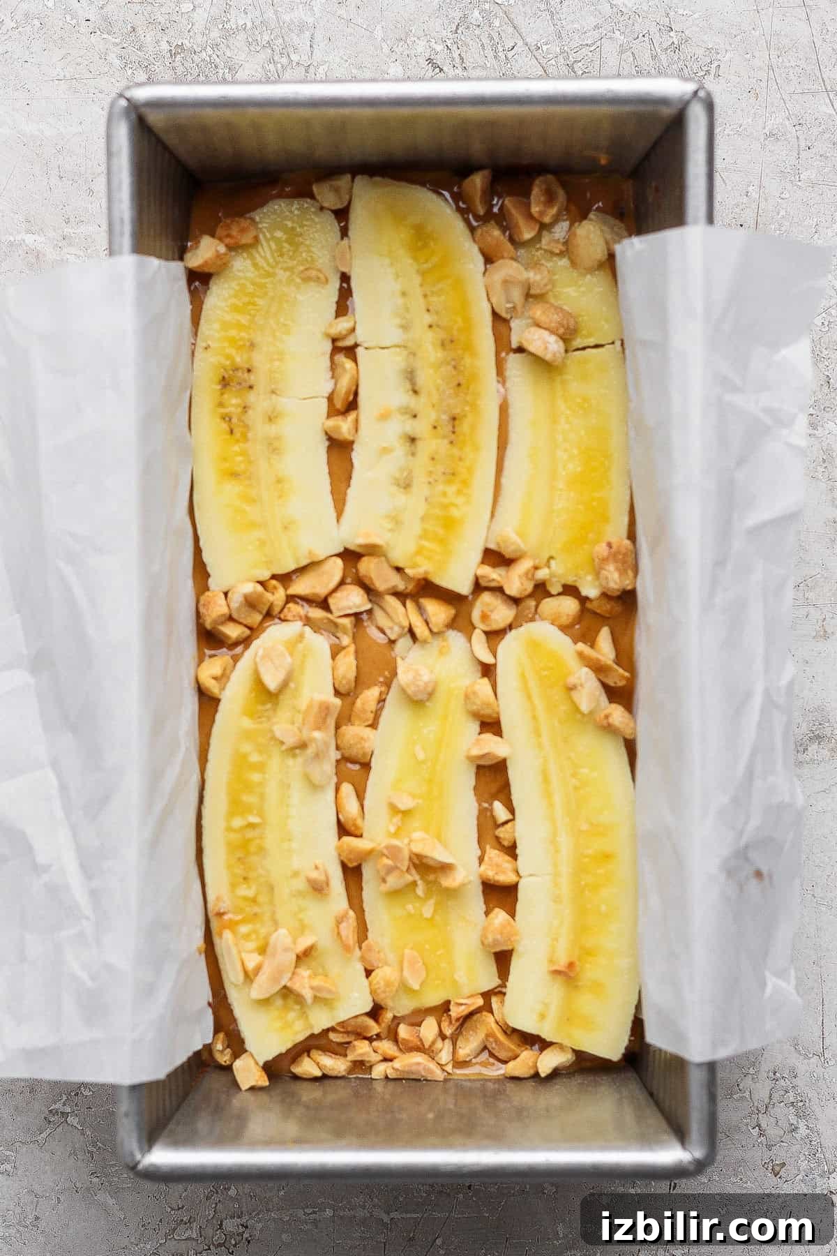 A baking tin lined with parchment paper, filled with batter, topped with halved banana slices and chopped peanuts.