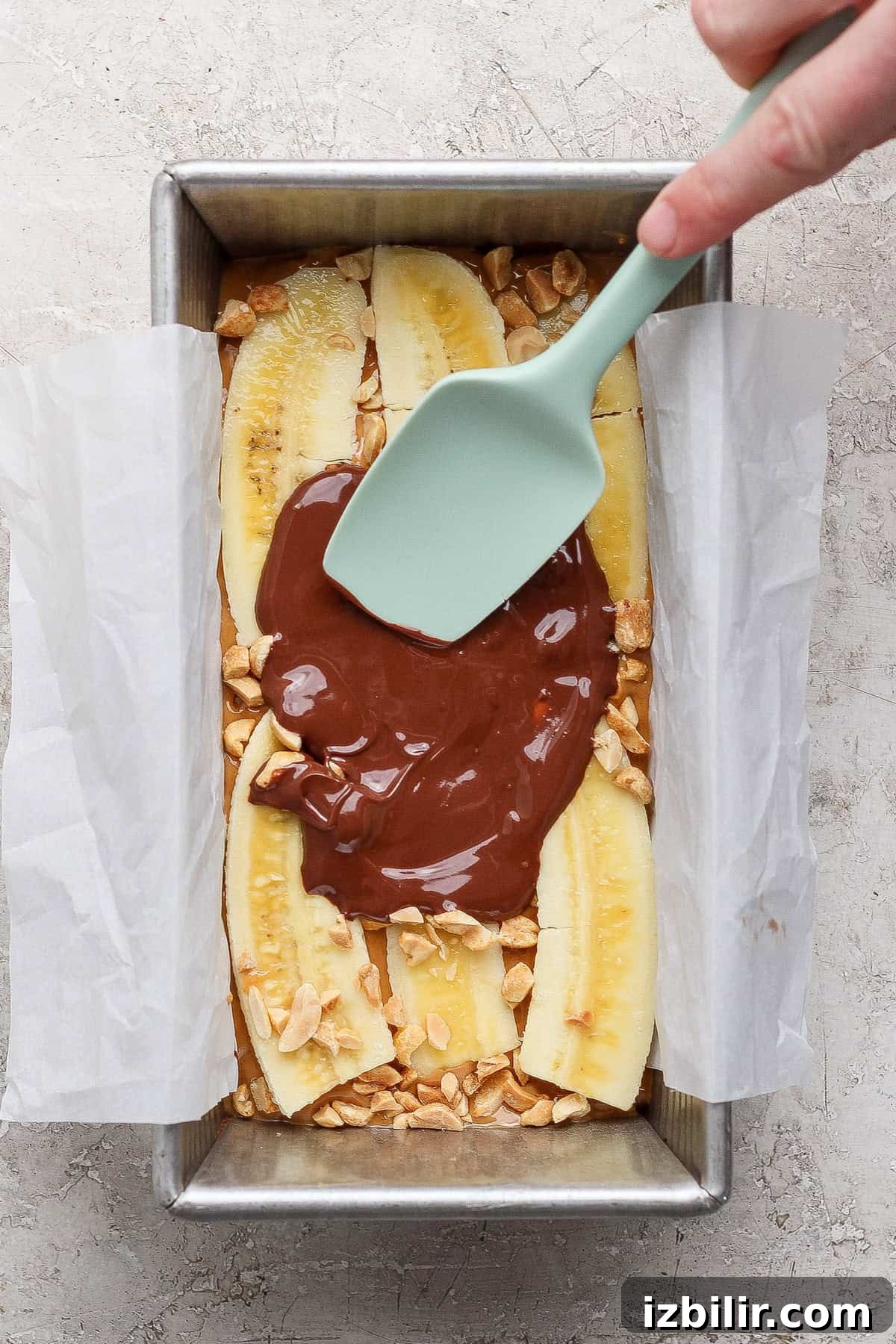 A hand spreads melted chocolate with a spatula over sliced bananas and chopped peanuts in a parchment-lined baking pan.